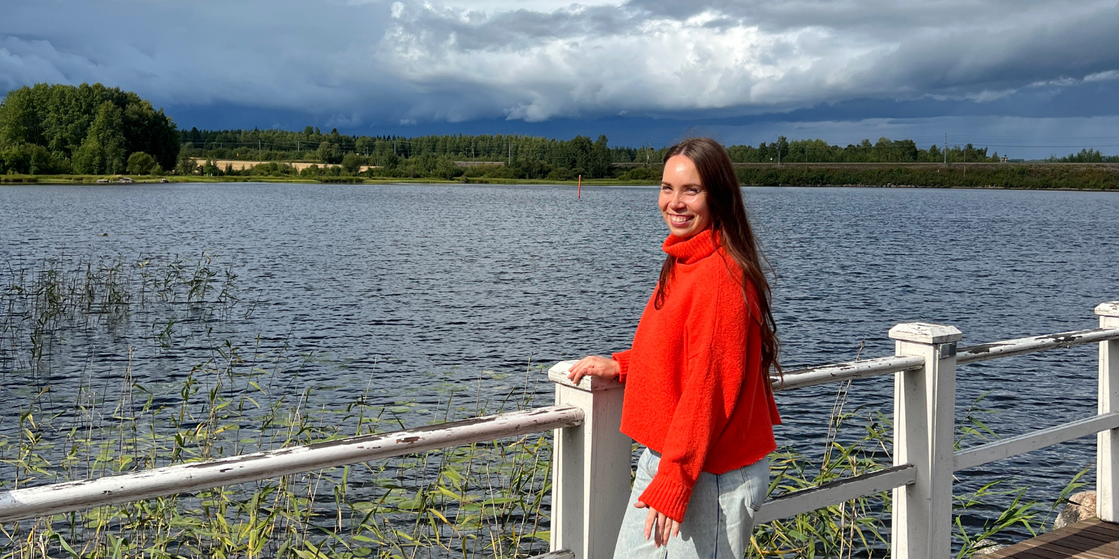 Nainen nojaa aitaan järven rannassa ja katsoo hymyillen kameraan