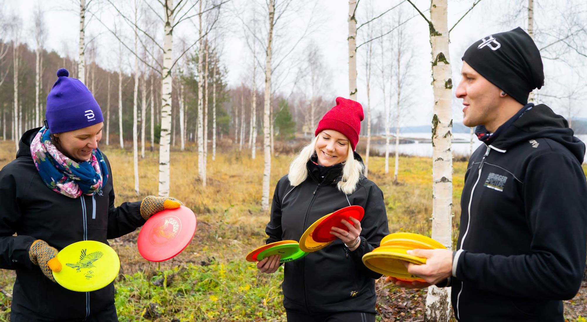 Kolme frisbeegolf-pelaaja koivikossa kiekot käsissään ja hymyssä suin.