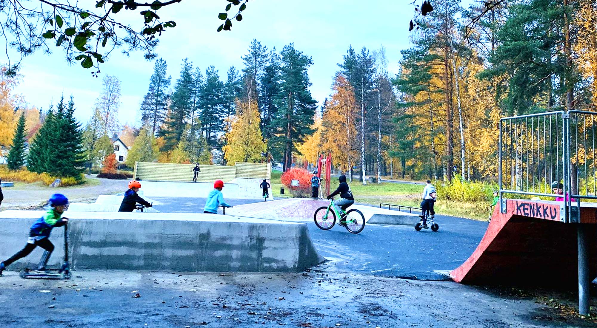 lapsila pyörillä ja scooteilla skeittipaikassa, ramppeja ja puiden reunustamana