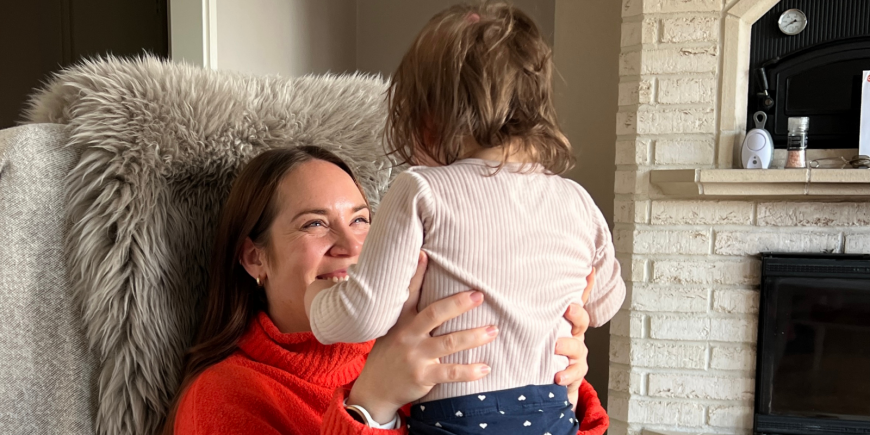 Nainen istuu nojatuolissa ja nostaa sylissään olevaan lasta. Lapsi on selin kameraan.
