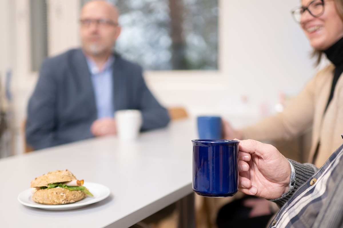 Joukko ihmisiä pöydän äärellä juomassa juomia kahvikupeista. Pöydällä on täytetty sämpylä. Ihmiset hymyilevät.