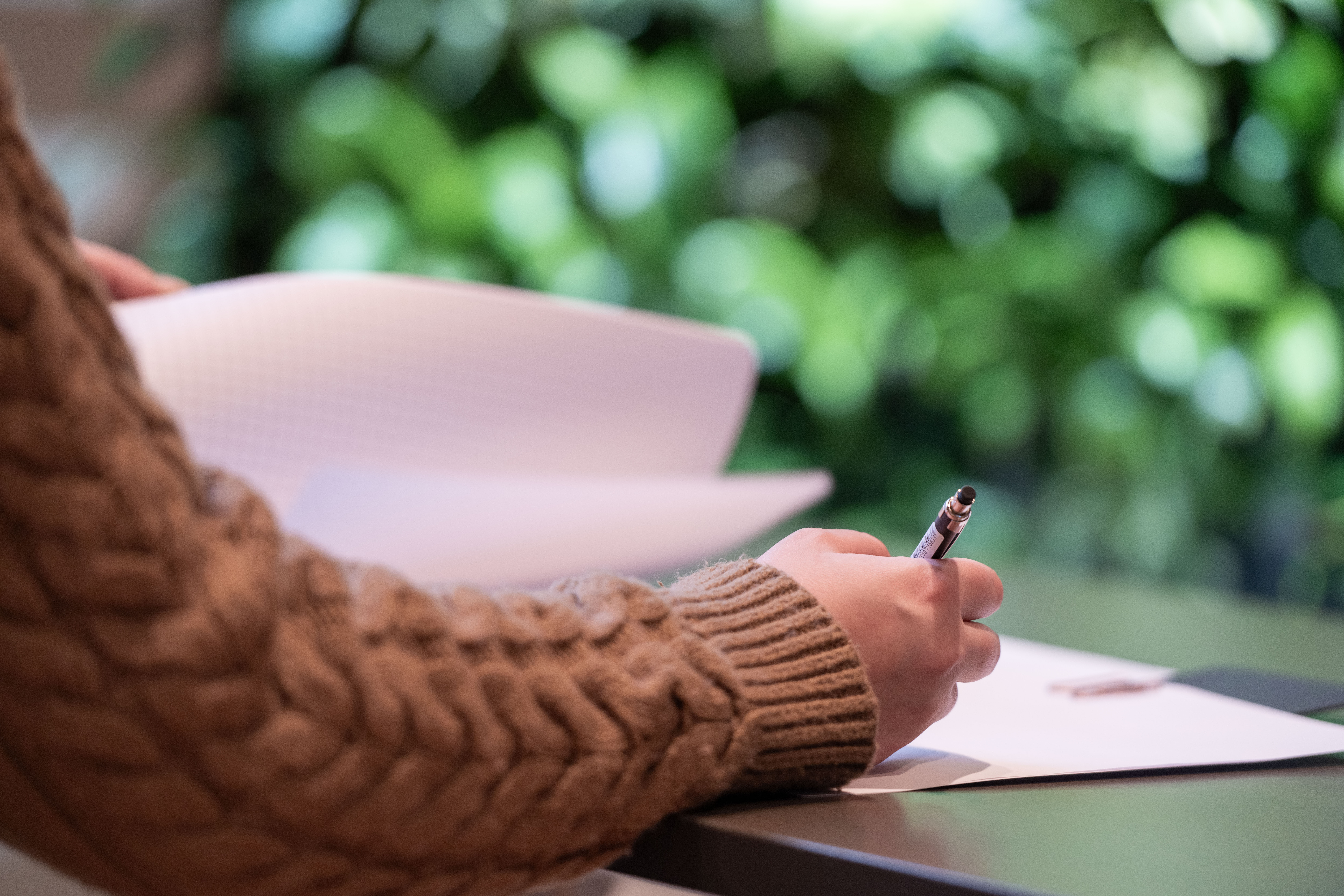 Ihmisen käsivarsi ja käsi. Kädessä on kynä ja vaikuttaa siltä, että käsi kirjoittaa jotain paperille, joka on käden alla. Taustalla näkyy sumeasti vihreitä kasvinlehtiä.