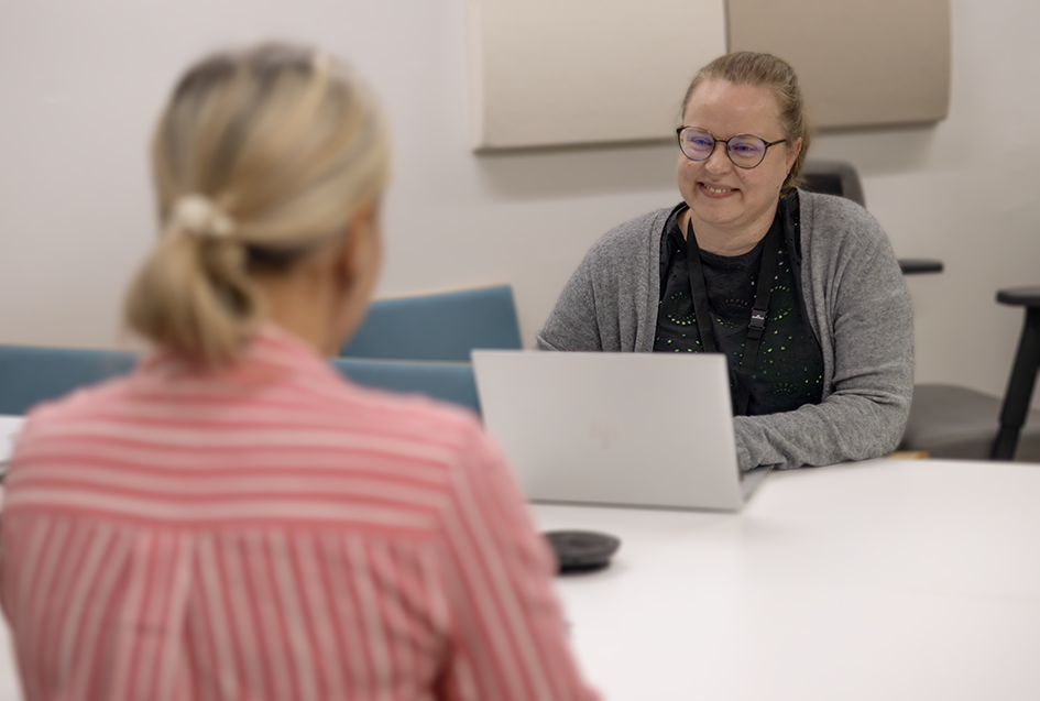 Työllisyysasiantuntija Liisa istuu pöydän toisella puolella ja hymyilee selin kameraan olevalle henkilölle, joka istuu vastakkaisella puolella pöytää.