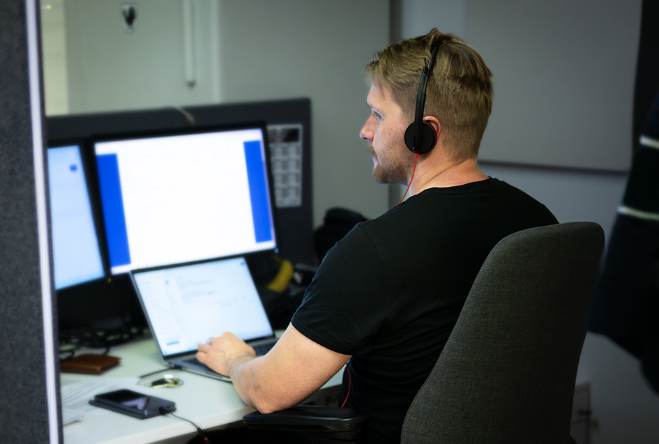 Mies istuu työpöydän äärellä ja katsoo tietokoneen ruutua. Hänellä on kuulokkeet korvilla. Pöydällä on kannettava tietokone ja kaksi näyttöä.